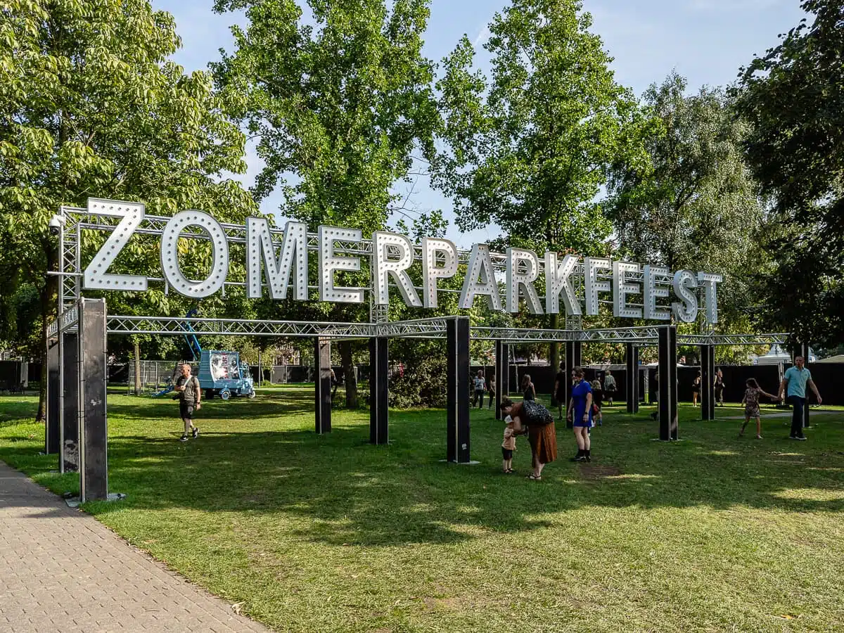 Große Metallbuchstaben bilden das Wort „ZOMERPARKFEEST“ auf einem Rahmen in einem grünen Park mit Bäumen in Venlo. Menschen, darunter Familien und Kinder, spazieren über den Rasen und entspannen sich dort. Es ist sonnig, und im Hintergrund hinter dem Schild ist ein blauer Transporter zu sehen.