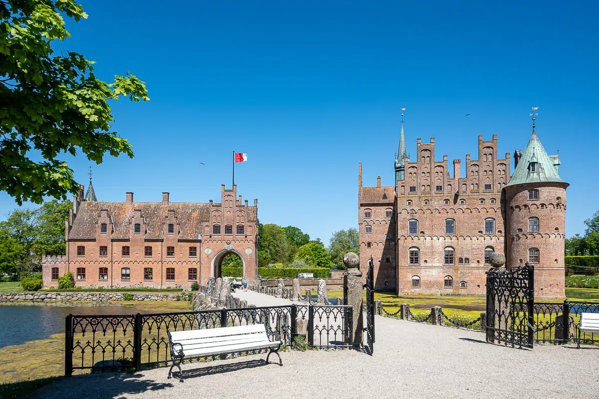 Ein heller, sonniger Tag am Schloss Egeskov in Dänemark mit seinen roten Backsteingebäuden, Türmen, Türmchen und einem Wassergraben. Ein Kiesweg führt über eine Brücke zum Eingang des Schlosses, mit weißen Bänken und einem verzierten schwarzen Geländer im Vordergrund. Über dem Tor weht die dänische Flagge.