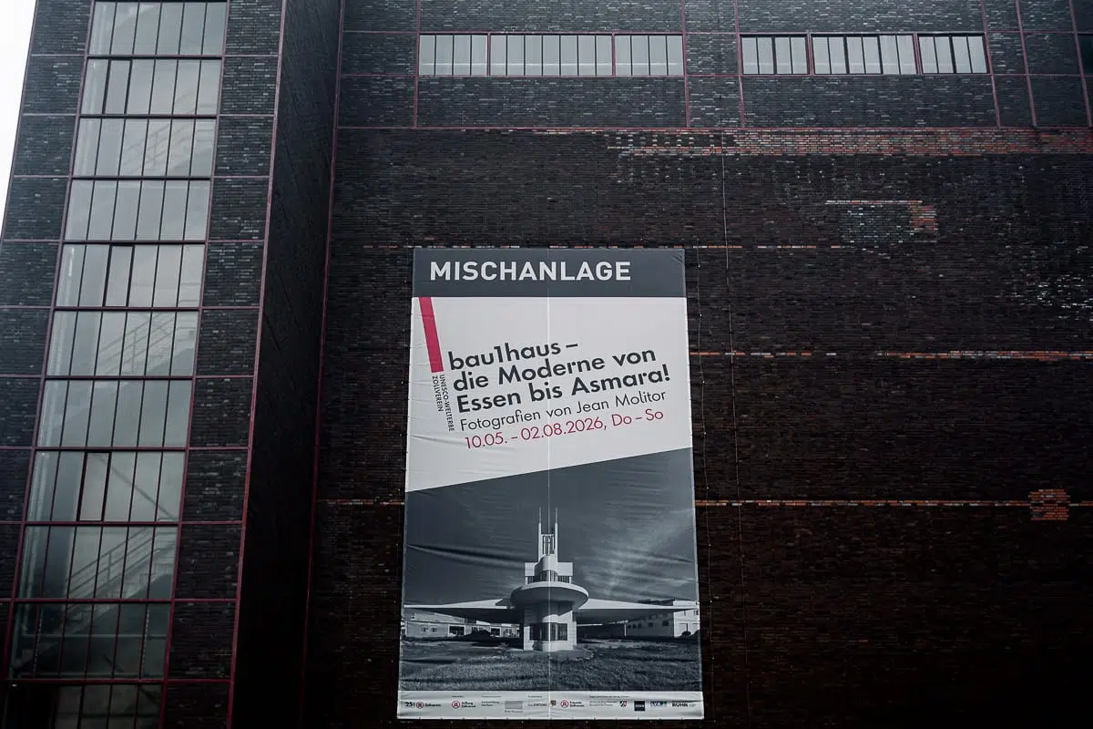 Auf einem großen Plakat an einem dunklen Backsteingebäude steht „MISCHANLAGE“ und wirbt für eine Bauhaus-Fotoausstellung von Jean Molitor, auf dem ein Schwarz-Weiß-Foto eines modernistischen Gebäudes zu sehen ist. Die Veranstaltungstermine und der Name des Fotografen sind angegeben. Links sind rote Fensterrahmen zu erkennen.